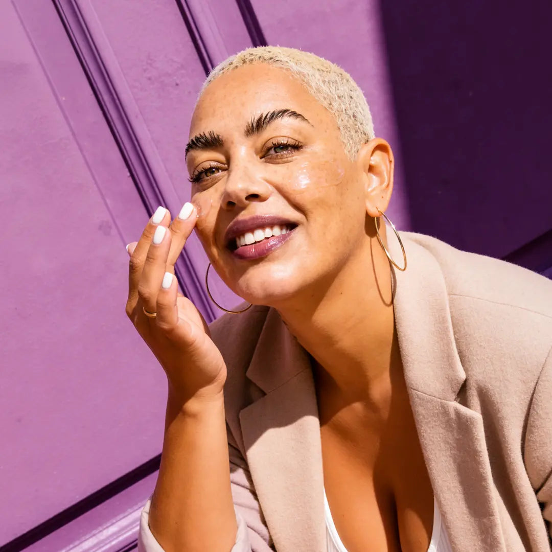 Smiling model applying Yelf Spot Off Vitamin-B3 Gel outdoors in bright sunlight with glowing skin, vibrant purple backdrop and fresh summer skincare aesthetic.