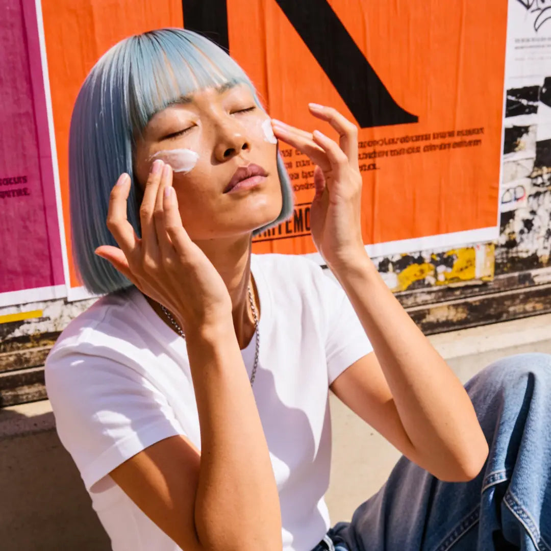 Model applying Yelf Aging Bye Bye Hyaluron Day Cream outdoors in bright sunlight with colorful street posters, creamy skincare texture and modern urban lifestyle mood.