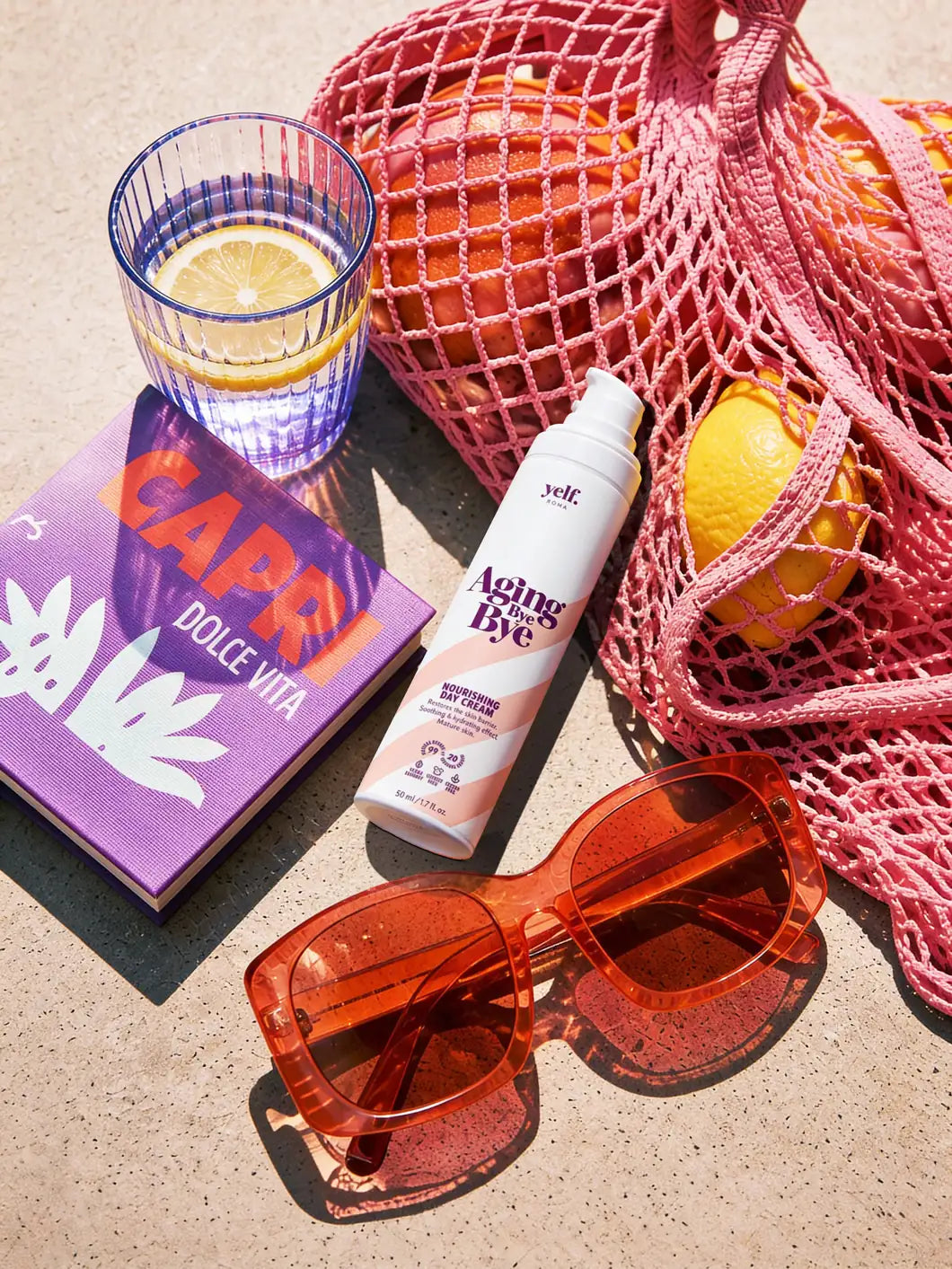 Yelf Aging Bye Bye Nourishing Day Cream with oranges in mesh bag, coral sunglasses and lemon water glass on concrete surface