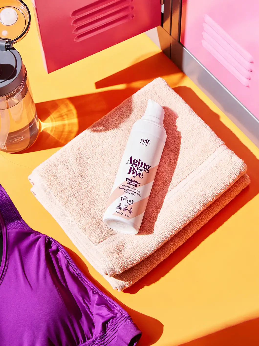 Top view of Yelf Aging Bye Bye Vitamin C Serum placed on a folded towel in a colorful gym locker setting with sports bottle, headphones and activewear in bright sunlight