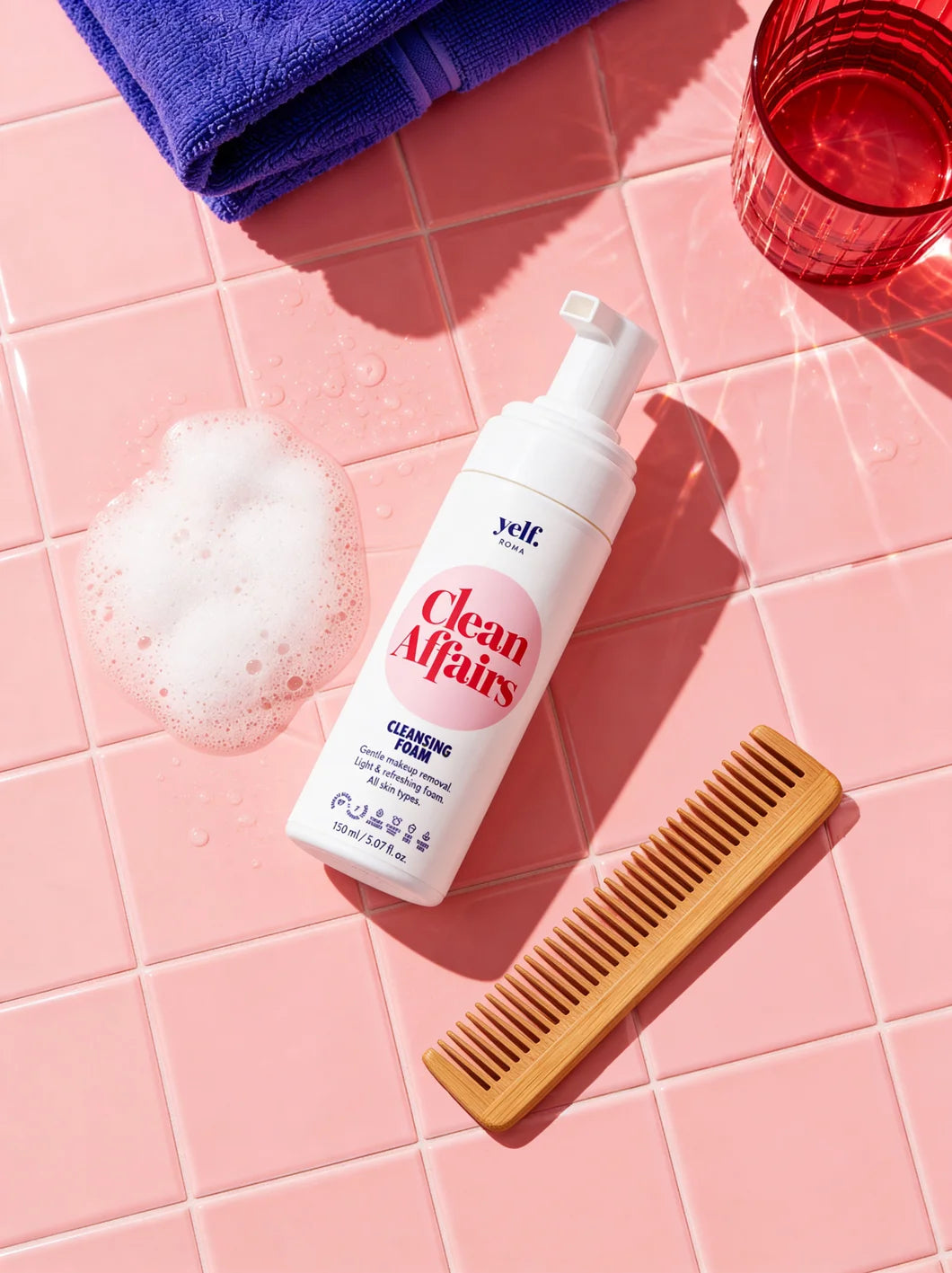 Yelf Clean Affairs Cleansing Foam on glossy pink bathroom tiles with soft foam, a red glass, towel and wooden comb in bright sunlight.