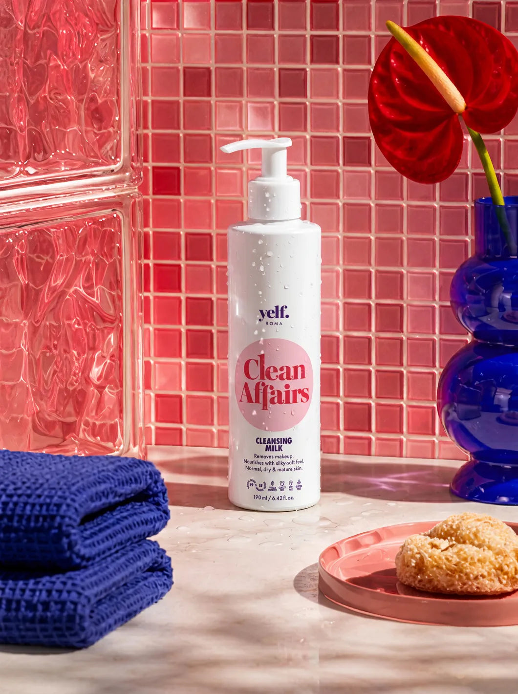 Yelf Clean Affairs Cleansing Milk standing on a marble bathroom counter with pink mosaic tiles, glass blocks, blue towels and bold red-blue decor in bright studio lighting.
