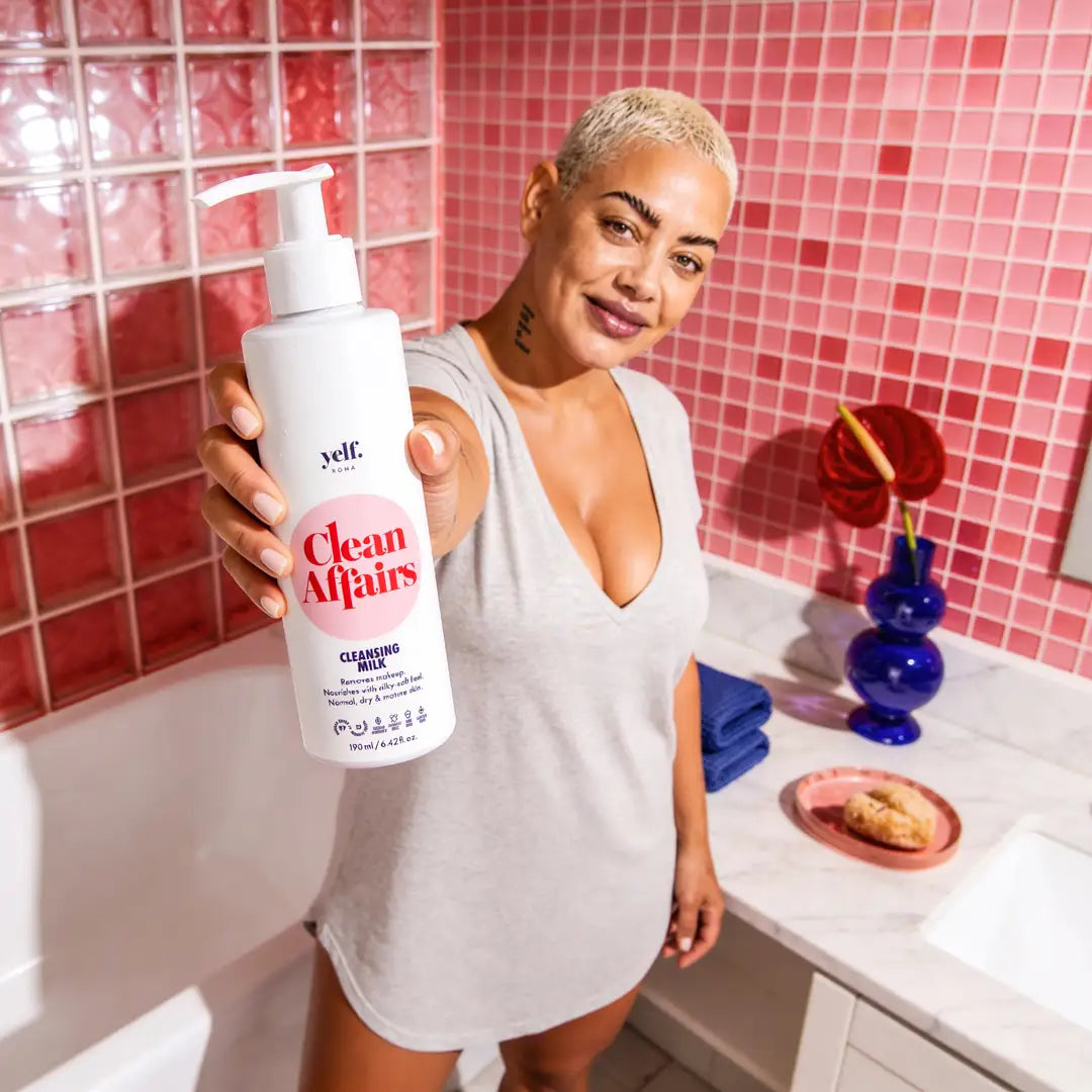 Smiling model holding Yelf Clean Affairs Cleansing Milk in a vibrant pink bathroom with glossy tiles, bold lighting and playful self-care atmosphere.