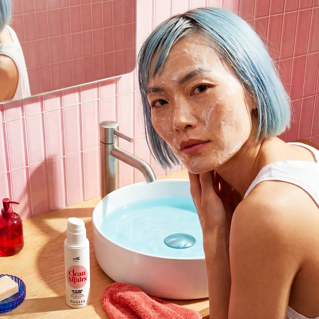 Model cleansing the face with Yelf Clean Affairs Oil-to-Milk Cleanser in a warm pink bathroom with soft skincare foam, glossy tiles and modern self-care atmosphere.