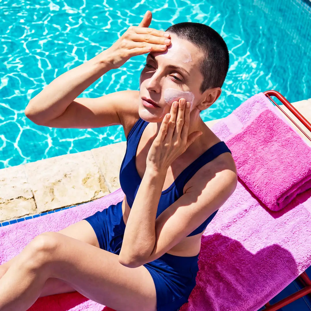 Woman applying Yelf Viva el Boost Hyaluron Cream while relaxing poolside on a pink sun lounger in bright summer sunlight.